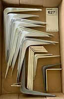 Multiple size metal 90-degree brackets stacked in a cardboard box, primarily white with a few gray brackets and mounting screws.