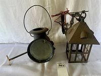 Close-up of two lanterns showing details: black carriage lamp with round glass lens and metal handle, and rusty hanging roofed lantern with glass panels and electrical mounting bracket, displayed against a plain white background.