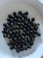 Top-down view showing a collection of about 77 black marbles with orange and yellow swirls inside a white bowl.