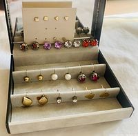 Wide view of 16 pairs of assorted stud earrings with different colored stones and shapes organized neatly in a grey jewelry organizer box.