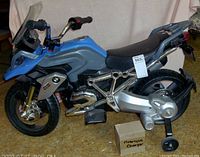 Side view of blue and grey electric ride-on BMW motorcycle with black seat and training wheels on carpeted floor, labeled charger box beside it.