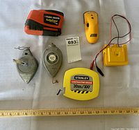 Photo showing the full set of six carpenter measuring tools arranged with a ruler for scale: two tape measures, two plumb bobs, one stud sensor, and one small electronic yellow tool with wires