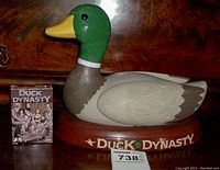 Photo showing the Duck Dynasty talking/singing duck figurine with visible articulation on head and feathers, alongside the boxed deck of Duck Dynasty playing cards.