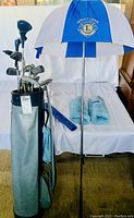 Full view showing golf bag holding multiple kids golf clubs, the blue and white Great Lakes Lions golf umbrella standing beside it, and two blue club head covers lying on a surface