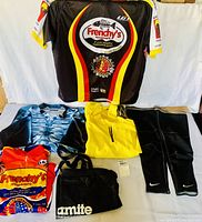Full lot laid out showing three men's cycling jerseys, black Nike knee sleeves, and a black Dynamite bag