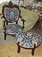 Front view of dark carved wooden armchair with blue and gold damask upholstery and matching round footstool with fringe trim.
