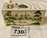 Front view of the ceramic holder with seven vintage hat pins inserted, detailed hand-painted green landscape artwork visible, showing trees, buildings, and water.