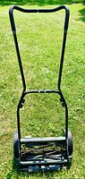 Front view of black metal frame reel mower with padded handle and cutting reel base on grass.