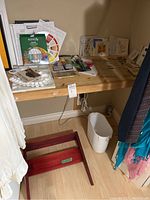 Overview of art supplies including paints, brushes, and greeting cards on a wood shelf with a trash bin below and a wooden item on the floor.