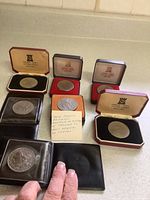 Six commemorative coins of Queen Elizabeth II in presentation boxes and plastic sleeves on a counter, some with colored velvet linings and a note about the reign of British coins.