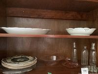 Shelf showing multiple vintage plates, glass bottles, and serving dishes in wood cabinet.