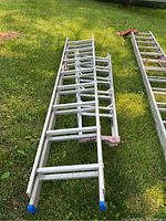 Two collapsed aluminum ladders side by side on grass, showing overall lengths and foot caps