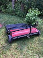 Red White Outdoor lawn sweeper with black handle and attached black fabric collection basket in grass area, showing wear and use.