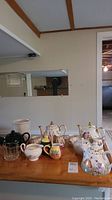 Wide view of all items including two Sadler teapot sets, creamers, sugar bowls, black wine bucket, and clear glass tumbler on wooden table.