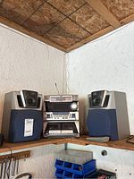 Full stereo setup on wooden corner shelf including two blue cloth speakers, central unit, and remote on shelf