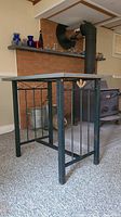 Table against wall showing metal base with vertical bars, decorative scrolls, and wicker leaf center accent. Painted wood tabletop on black metal frame.