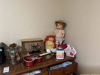 Full view of the lot on a wooden dresser including toy sewing machine in box, jar of painted gold pinecones, red ukulele, two dolls, Stack Attack game and a decoration kit.