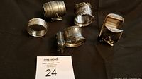 Lot of 5 assorted antique silver plate napkin rings displayed on black fabric with auction tag