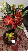 Full view of artificial flower arrangement in black leatherette vase and wire bowl with decorative fruits on red patterned carpet.