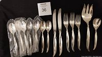 View of arranged flatware pieces showing forks, knives, spoons including some wrapped in plastic; highlights total quantity and condition with minor tarnish visible on some pieces.