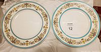 Two round plates with decorative floral and scrolling ornamental borders accented in blue and gold bands, placed on white cloth background.