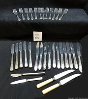 Overview of silver plated forks on top row in set of 12, with butter knives and spreaders below, some with bakelite handles.
