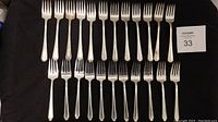 Two sets of silver forks laid out on black fabric: 12 Rogers & Sons "Paris" forks top row, 11 1847 Rogers "Legacy" forks below.