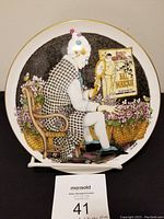 Full front view of the Royal Doulton Make Me Laugh limited edition plate showing character seated on a chair with flowers and gold trim.