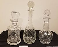 Three different patterned crystal decanters standing side by side on a black cloth against a white wall. Shows clear detail of design and shape of decanters and stoppers.