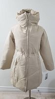 Cream puffy coat with a high collar, zipper closure, drawstring at waist, and long sleeves displayed on a dress form with tags attached.