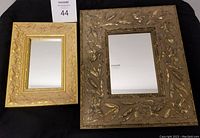 Both mirrors shown side by side on a dark cloth background. Smaller mirror on left, larger on right. Clear reflective glass and ornate gold-tone frames with intricate floral and leaf embossing.