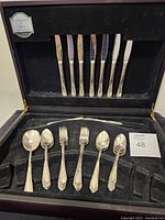 Closed wooden case containing 7 knives in upper lid and lower section with spoons and forks arranged in slots, showing "Meadowbrook Heather" pattern handles and some wear marks on flatware and case
