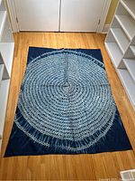 Full view of rectangular indigo and white tie-dyed textile with concentric circular and linear star burst patterns laid flat on wooden floor.