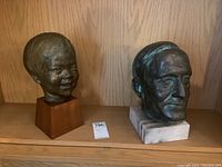 Two busts displayed side by side on a wooden shelf showing their bases, one man on marble base and one child on wood base