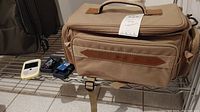 Photo of beige Optex camera bag with brown leather accent, BlackBerry phone, several battery chargers, and a small calculator on metal rack.