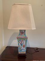 Front view of the lamp showing ceramic body with paintings and beige fabric shade on wood base