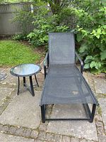 Photo showing black folding lounger with metal frame and matching round glass top side table on outdoor stone patio with greenery background.