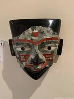 Front view of the black porcelain mask with multicolored stone mosaic, red painted decorative accents, hollow eyes, and flat top with ear pieces.