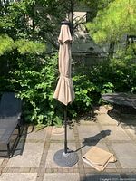 Patio umbrella closed, showing metal base, black metal pole, and tan folded fabric canopy.