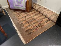 Area rug partially rolled and laid out on floor showing geometric patterns and solid brown border with fringe on one side.