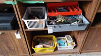 Overall view of cabinet shelf holding various tools, plastic baskets with miscellaneous hardware, and tray with hand tools and small parts.