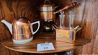 Photo of a copper teapot with white ceramic handle and spout, a copper oil and vinegar set with glass bottles and copper carrier stand, and copper fondue pot with brass accents on wooden shelf.