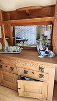 View of the oak craftsman hutch's top and middle sections showing built-in mirror, brass handles, carved floral details, and staged ceramic and glass decor items on the surface.