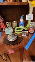 Decorative items arranged on a wooden cabinet shelf including beaded nesting boxes, gnome figurine, glass containers, candle jar, metallic sculptures, flower vase, and glass plate with spherical object.