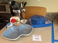 Photo showing assortment of kitchen items including blue enamel cookware set, utensils in metal holder, wooden cutting board, ceramic dishes, and floral-themed napkin holder
