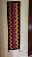Full view of the rectangular textile wall hanging with geometric zigzag pattern in black, orange, red, and blue, hanging on a wall.