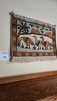 View of wool wall rug hung on wall showing colorful duck motif with natural scene including palm tree, mountains, and plants with fringed edges