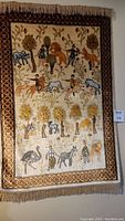 Full view of the animal-themed wall rug with fringe on top and bottom, showcasing foliage, animal, and human figure patterns with a geometric border.