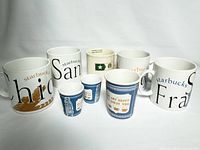 Photo showing four Starbucks 1990s city collector mugs: Orange County, San Francisco, Chicago, San Diego, alongside Starbucks Veterans mug and three blue Kikkerland ceramic NYC takeaway-style cups; two small and one large, arranged on white background.