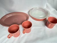 Photo showing three pink Fiesta mugs, pink Fiesta oval platter, and solid pink Pyrex casserole dish with clear lid.